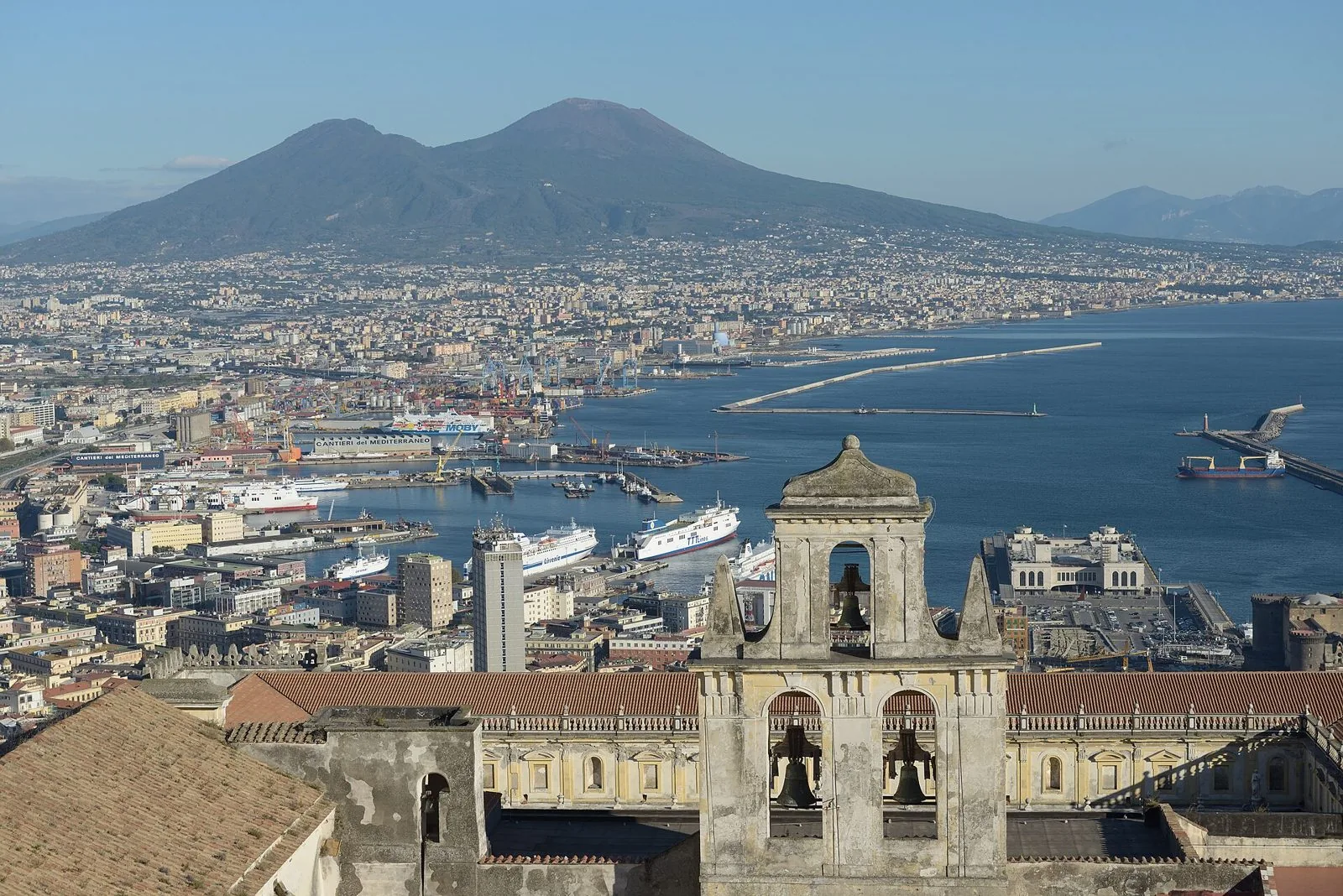 Panorama di Napoli dall'alto con il porto, il mare del golfo e il Vesuvio sullo sfondo, in primo piano il campanile e i tetti della Certosa di San Martino