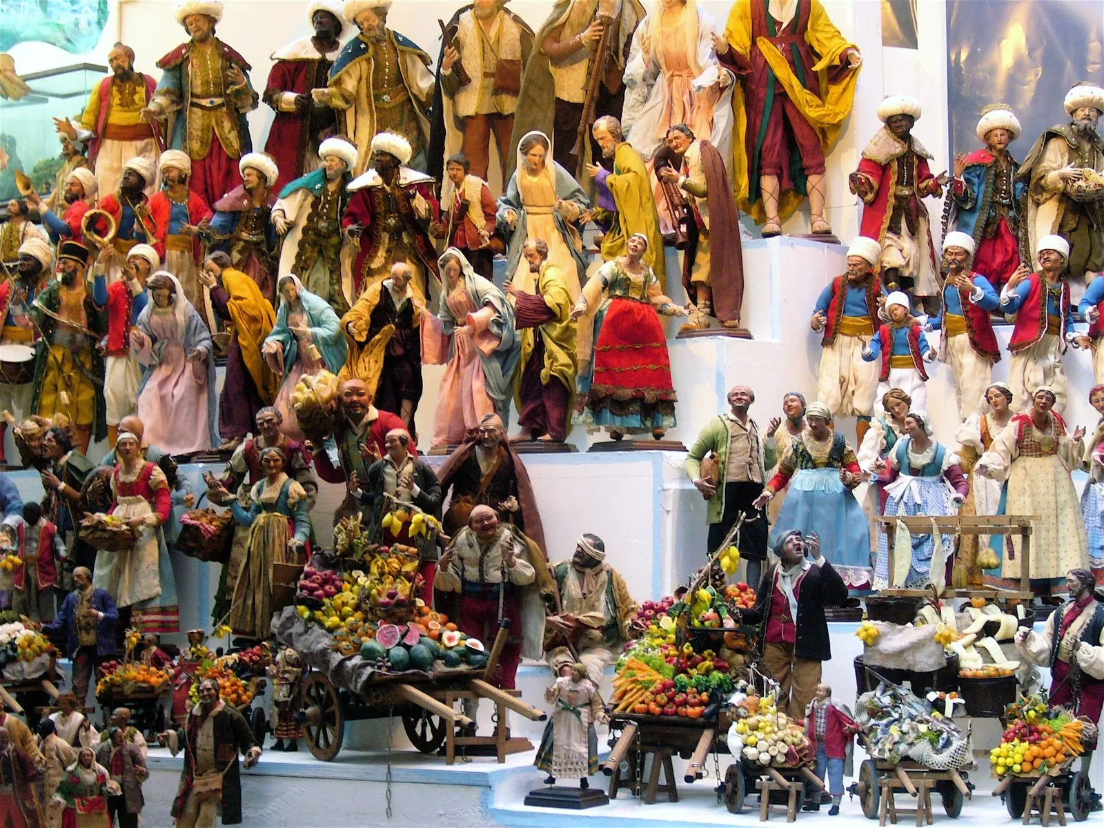 Pastori del presepe napoletano in terracotta e stoffa esposti a gradoni, con carretti di frutta e verdura in miniatura