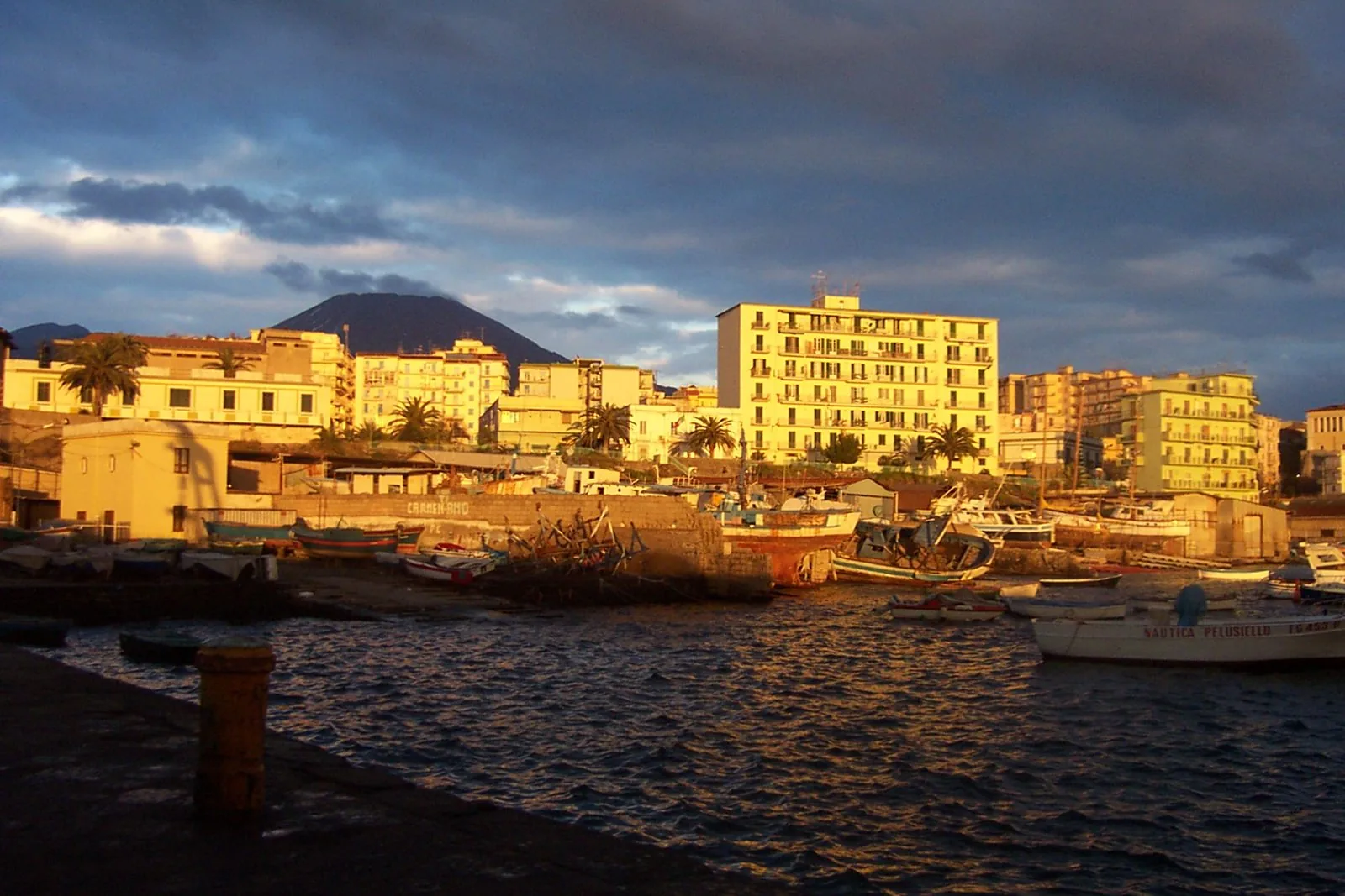 Il porto di Torre del Greco con barche ormeggiate e palazzi illuminati dal sole del tramonto, con il Vesuvio sullo sfondo